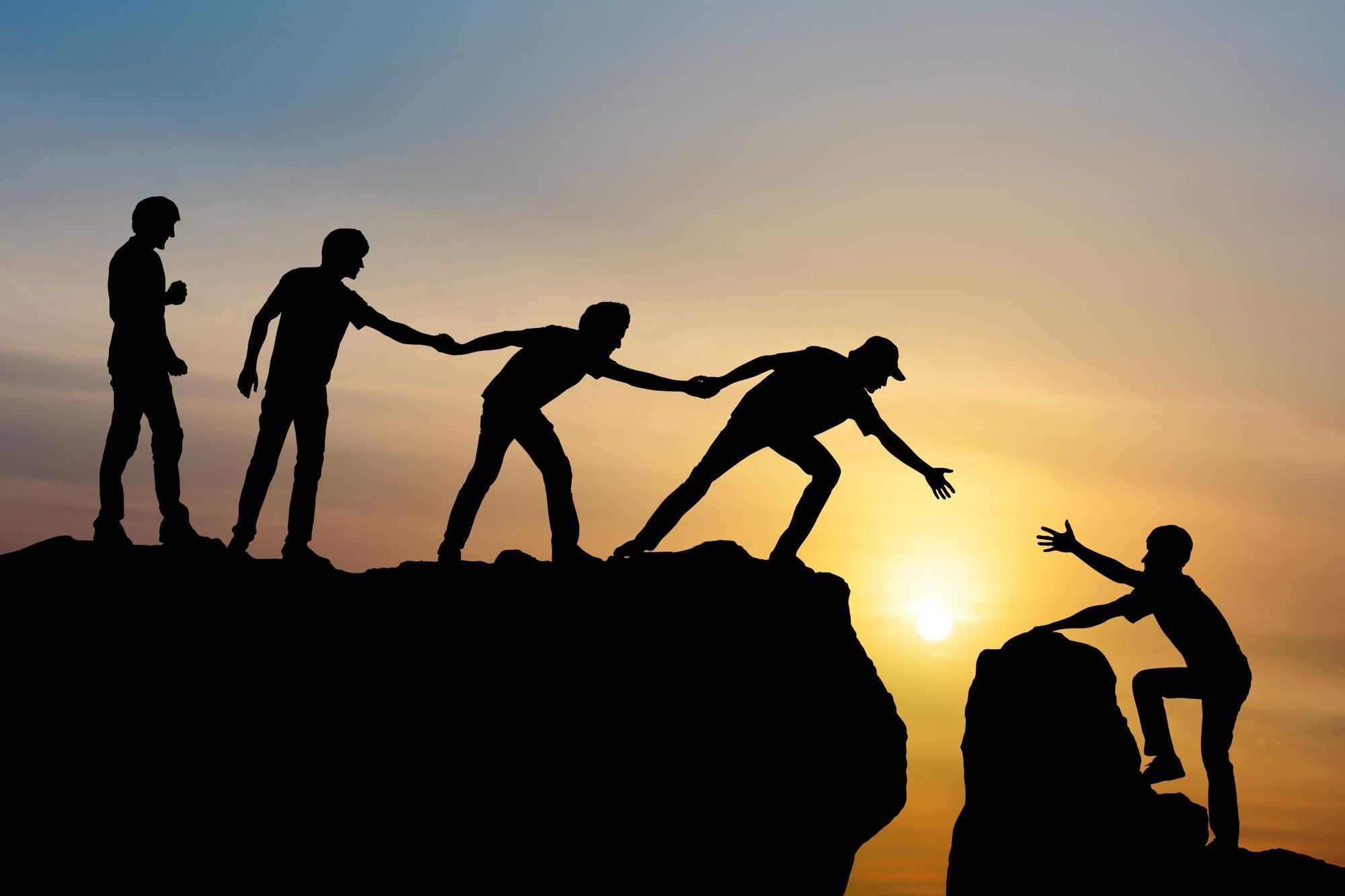 A group of silhouetted people help shine a climbing silhouette over the edge of a cliff with the sun setting in the backgr...